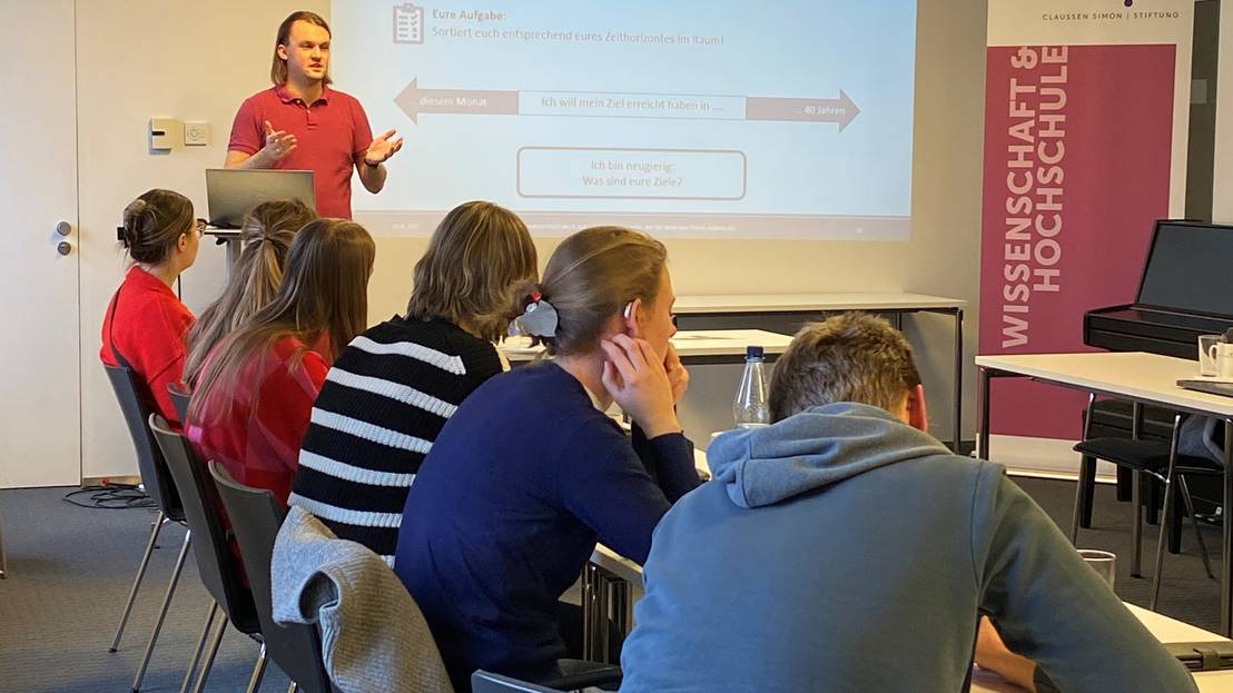 Ein Mann gibt eine Präsentation für sechs Teilnehmende, die an einem langen Tisch in einem Seminarraum sitzen.