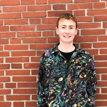 Porträt des Komponisten Dominic Wills in einem bunt gemusterten Hemd, vor einer gemauerten Wand stehend