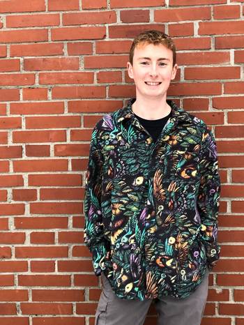 Porträt des Komponisten Dominic Wills in einem bunt gemusterten Hemd, vor einer gemauerten Wand stehend