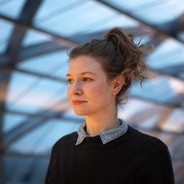 Foto einer Frau mit hochgesteckten Haaren, die links aus dem Bild raus schaut. Im Hintergrund eine Architektur aus Glas.