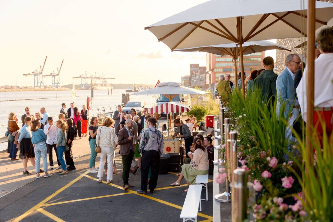 Foto von der der Fläche vor der Stiftung im Abendlicht. Es stehen viele Menschen da und unterhalten sich. Rechts sind Sonnenschirme zu sehen. Alles ist in goldenes Licht getaucht.