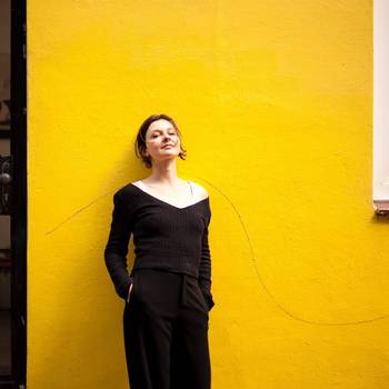 Porträt der Künstlerin Anne Pretzsch, in einem schwarzen Kleid vor einer gelben Wand lehnend