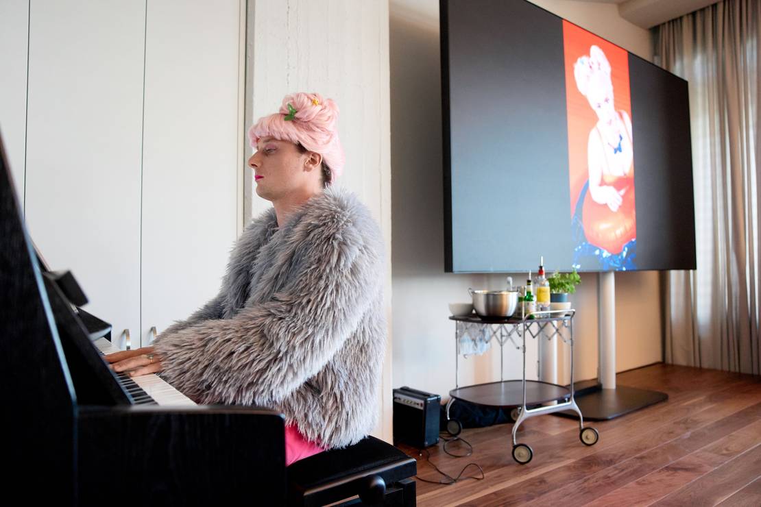 Ein Mann sitzt am Klavier und spielt. Er trägt eine rosa Perücke und einen Pelzmantel.