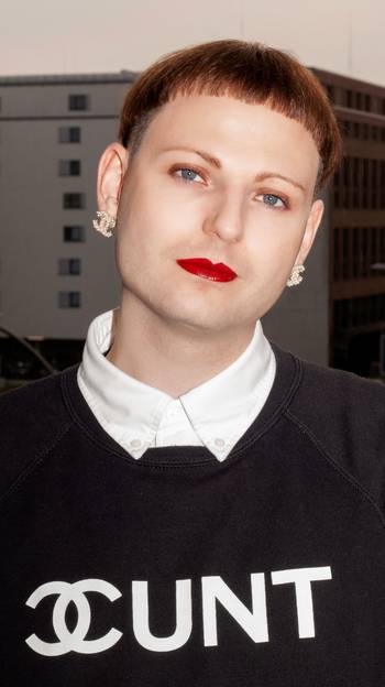 Porträt von René*e Reith, er trägt einen schwarzen Pullover mit weißer Aufschrift CUNT und das Chanel-Logo imitierend