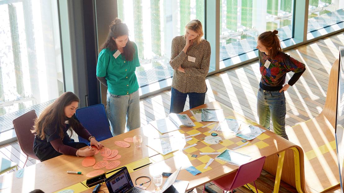 Vier Frauen in Gruppenarbeit an Tisch mit Präsentations-Materialien. Drei stehend und eine sitzend.