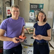 Foto von einem Mann und einer Frau in einer Werkstatt. Der Mann hält zwei Handys in der Hand. Beide lächeln in die Kamera.