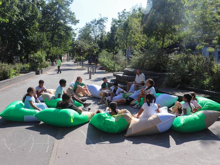 Eine Gruppe Kinder sitzt in der Sonne im Kreis auf großen grünen Kissen